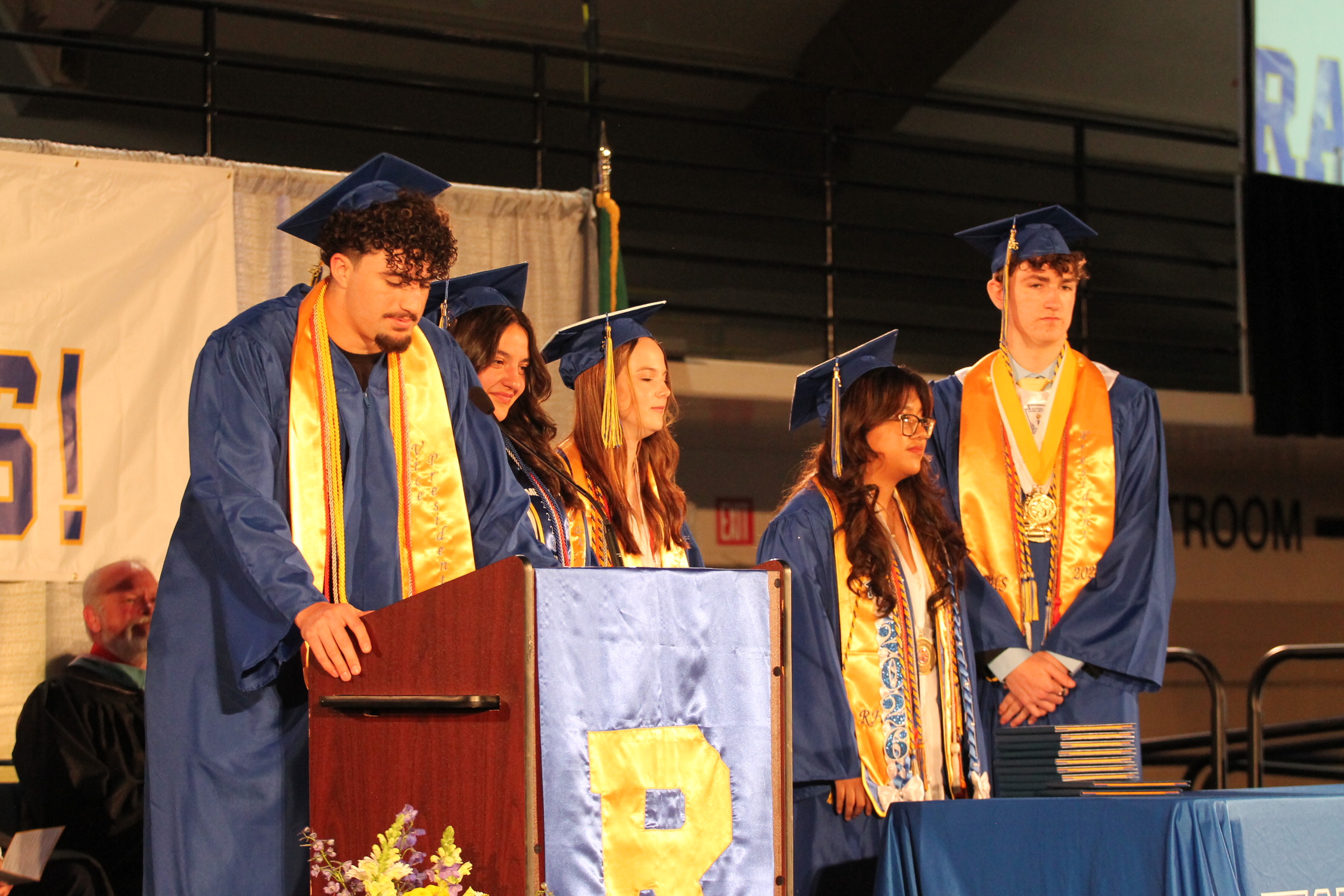 Five students in blue caps and gowns stand at a podium on stage, with one speaking.