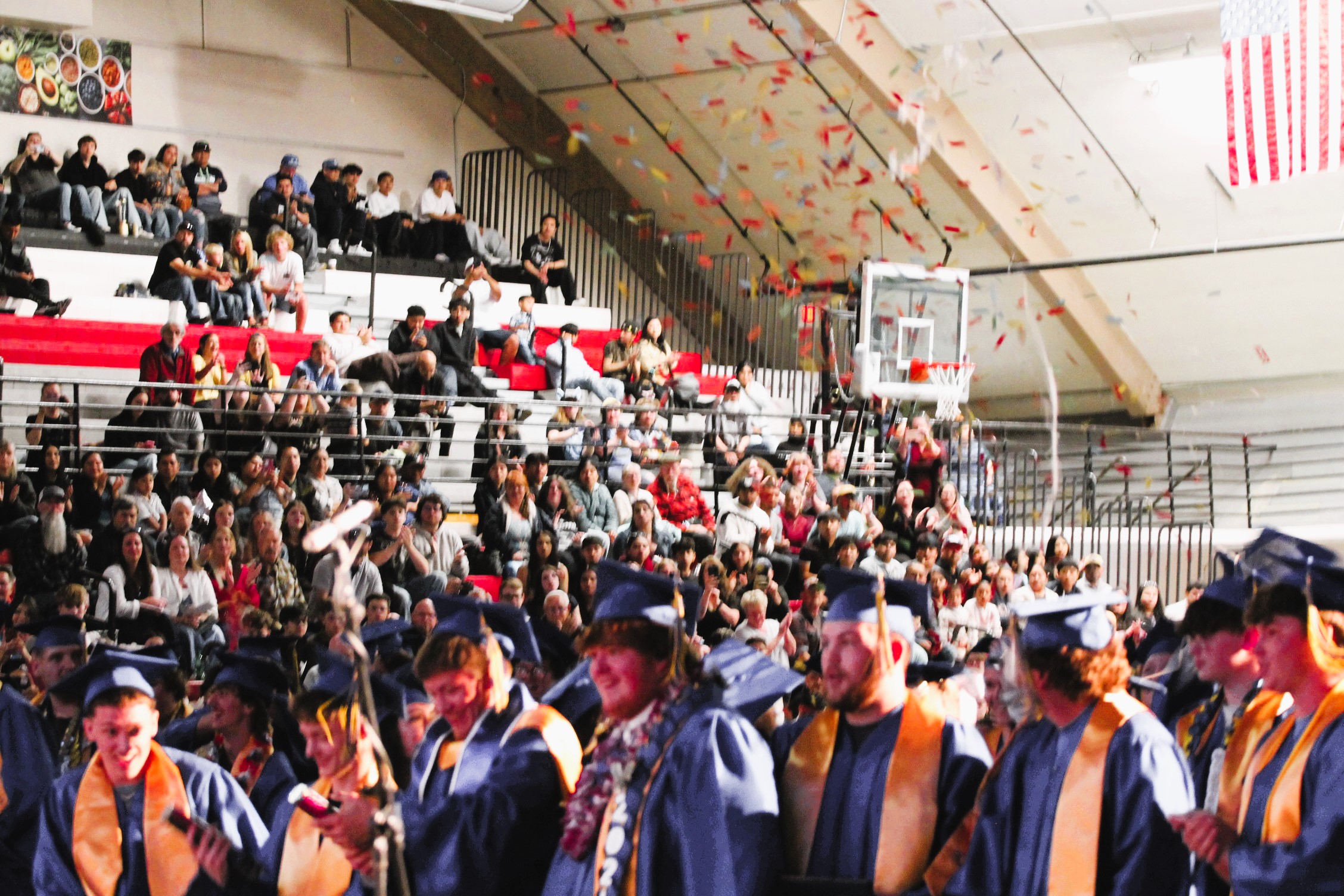 A large crowd of people in caps and gowns in a gymnasium with a basketball hoop.