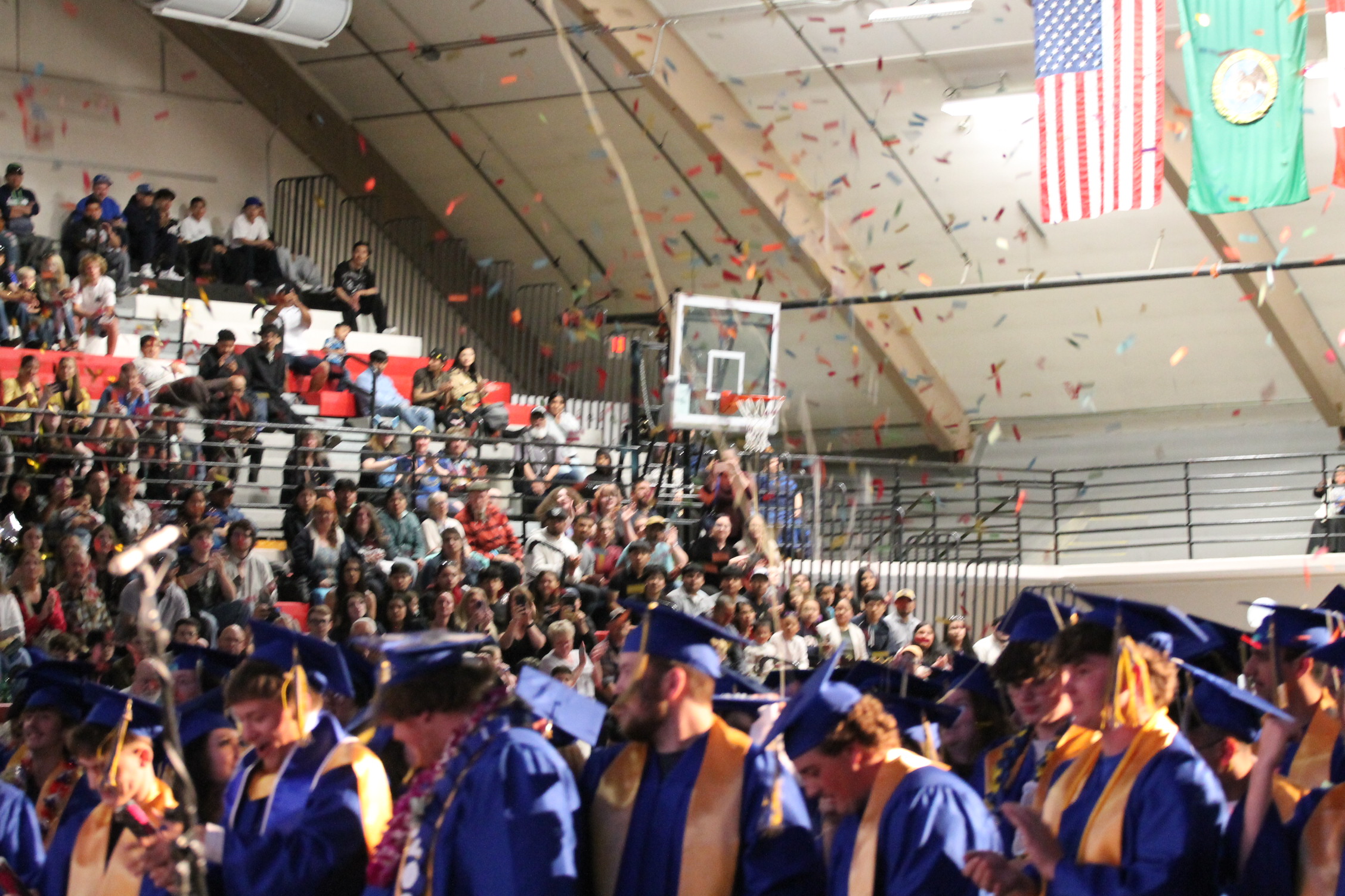 A graduation ceremony with graduates in blue gowns and caps, a basketball hoop in the background.