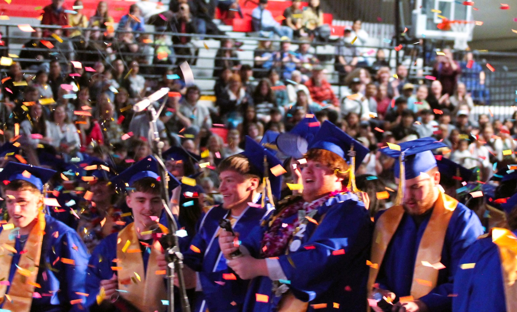 Graduates in blue gowns and caps, surrounded by confetti, stand in a stadium as an audience watches.