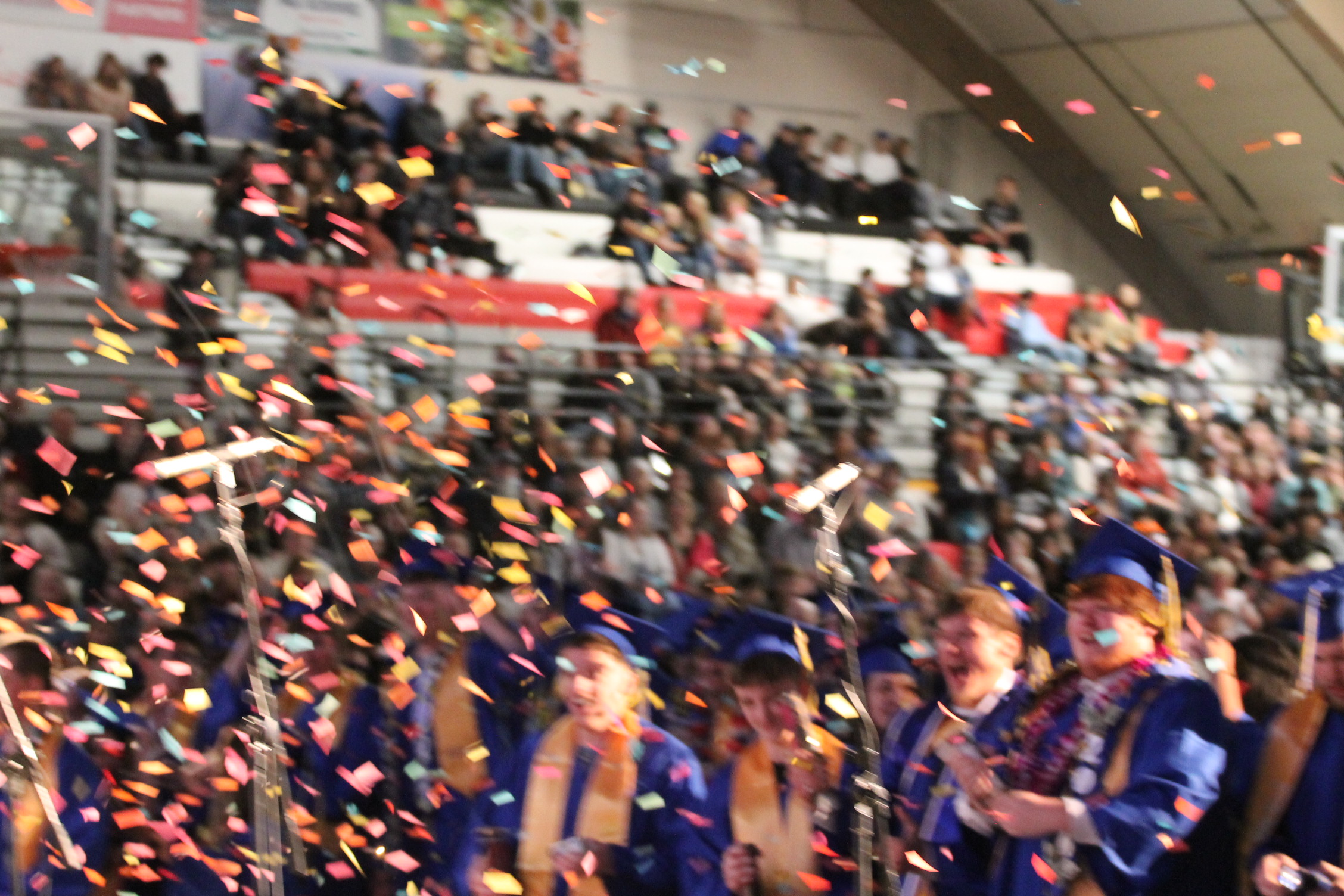 Graduates in blue gowns and caps in an indoor venue with multicolored confetti falling, and a large audience seated in bleachers.