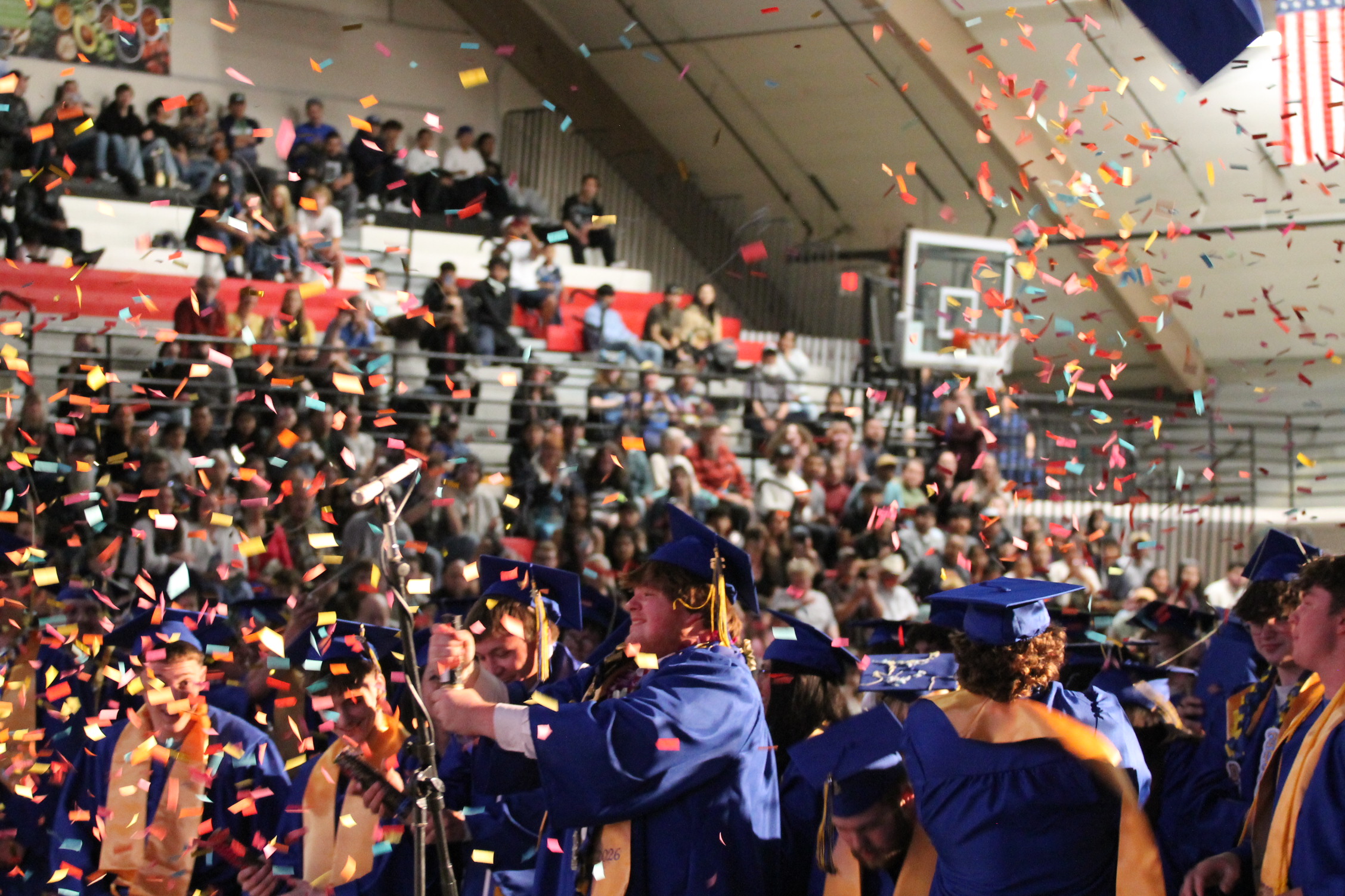A sports hall filled with people celebrating; confetti falls from above as graduates in blue gowns and hats celebrate.