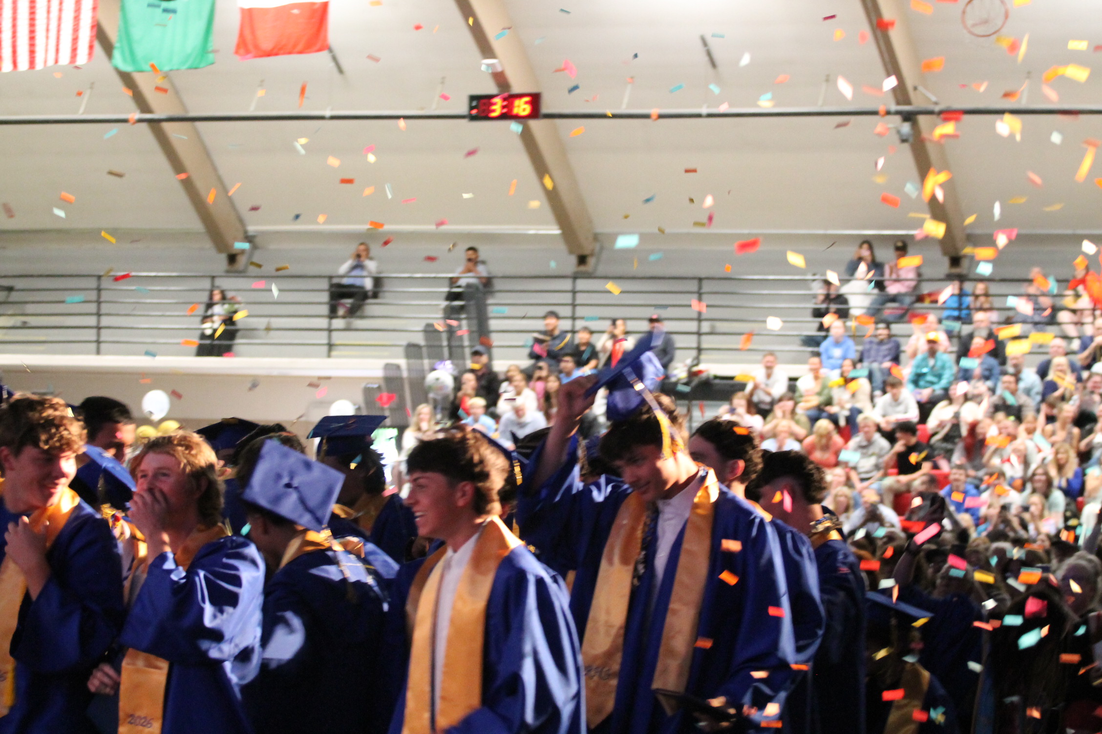 A group of graduates in blue gowns and hats celebrate in an auditorium. Confetti falls around them.