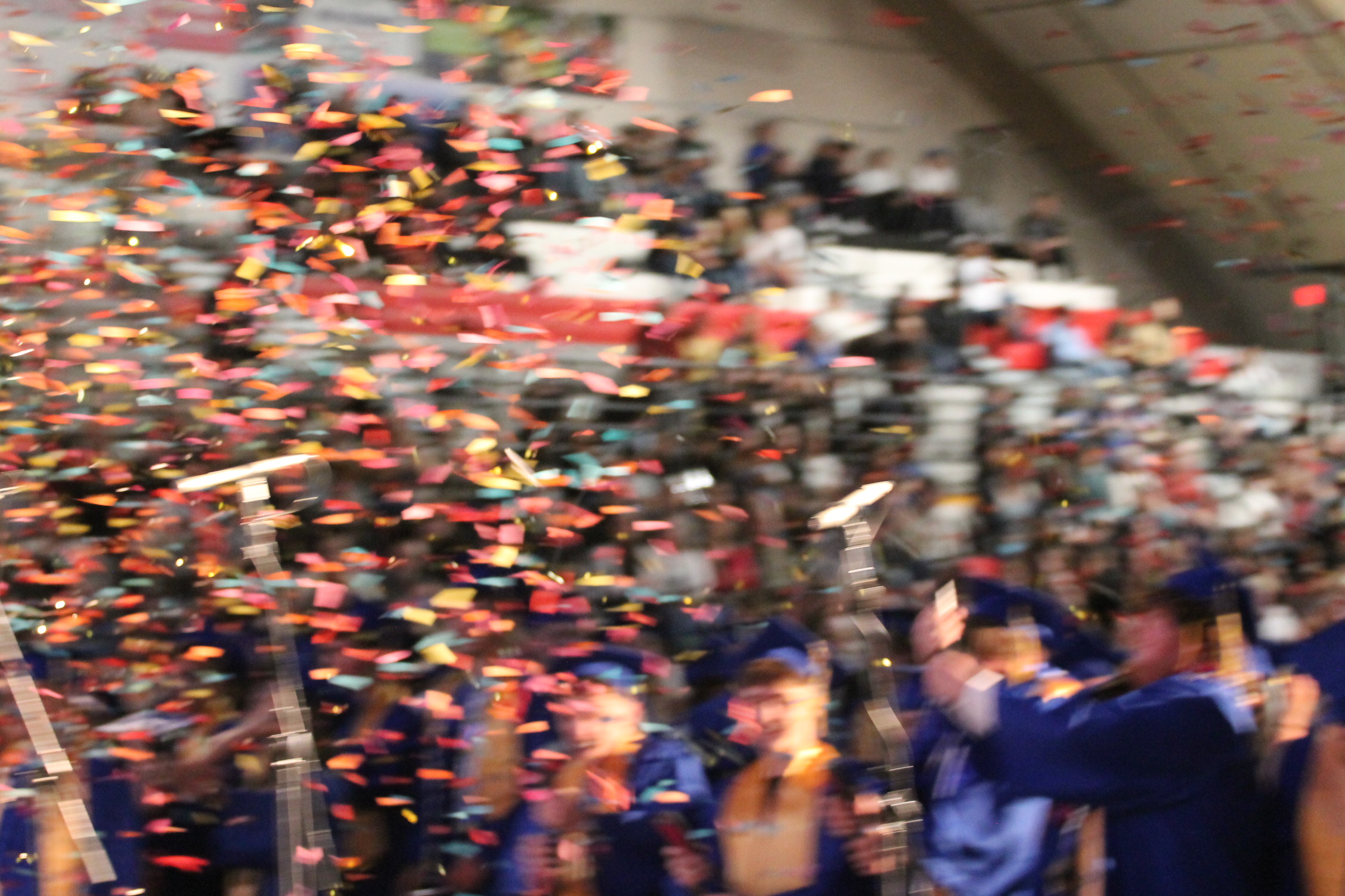 Graduates in caps and gowns celebrate with confetti in a large indoor venue. Audience members sit on bleachers.