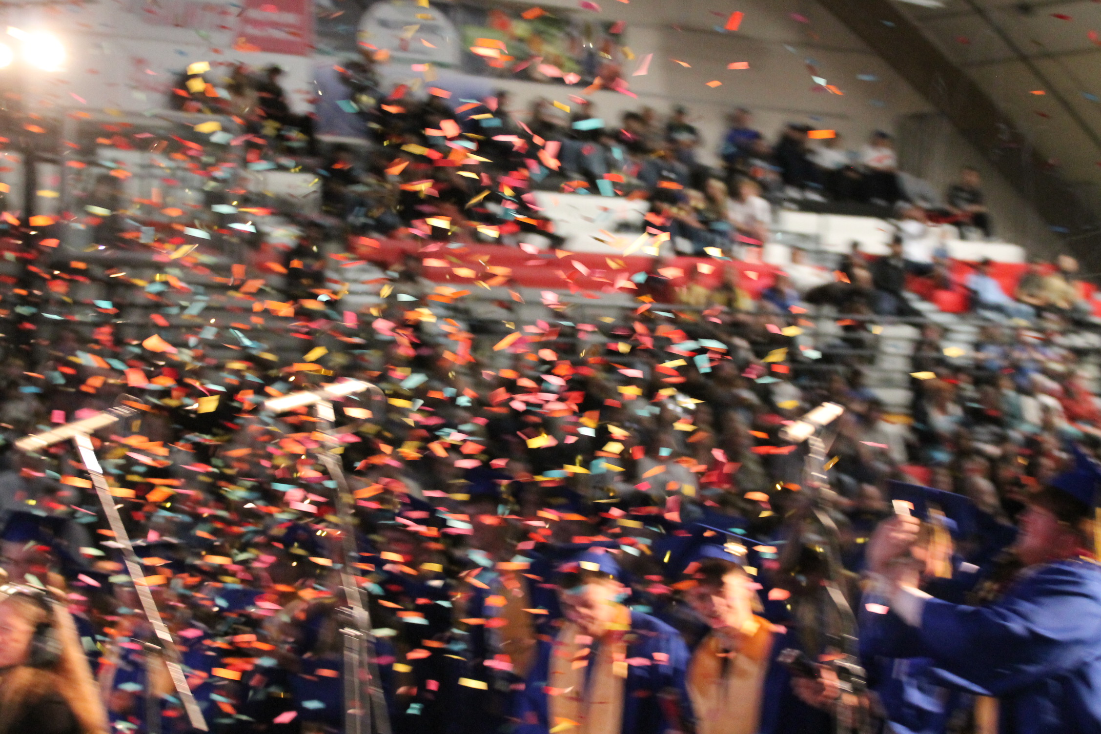 Confetti falling at a graduation ceremony with people in blue gowns and caps, some holding cameras.