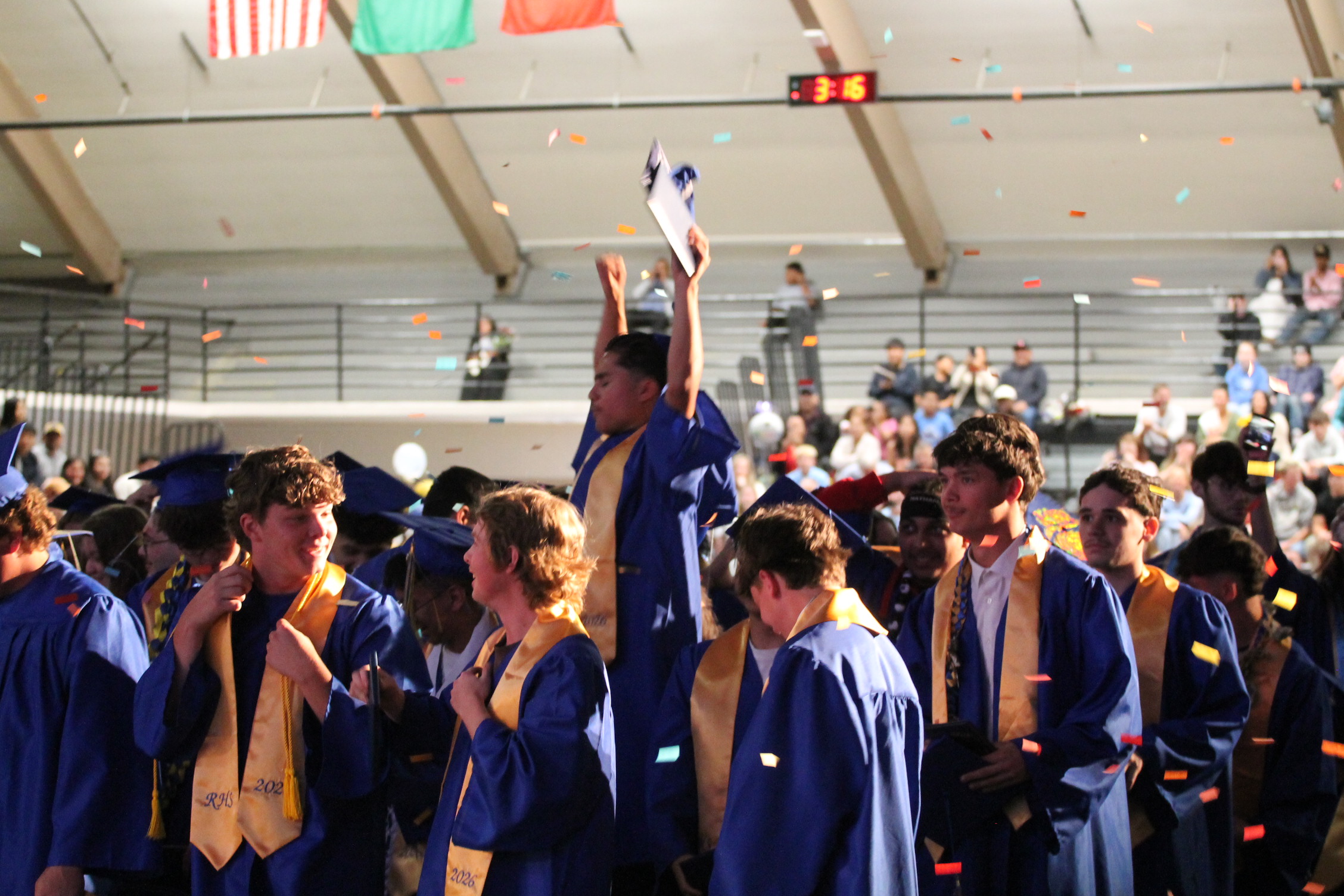 Graduates in blue gowns and caps celebrating at a ceremony with confetti and spectators in the background.