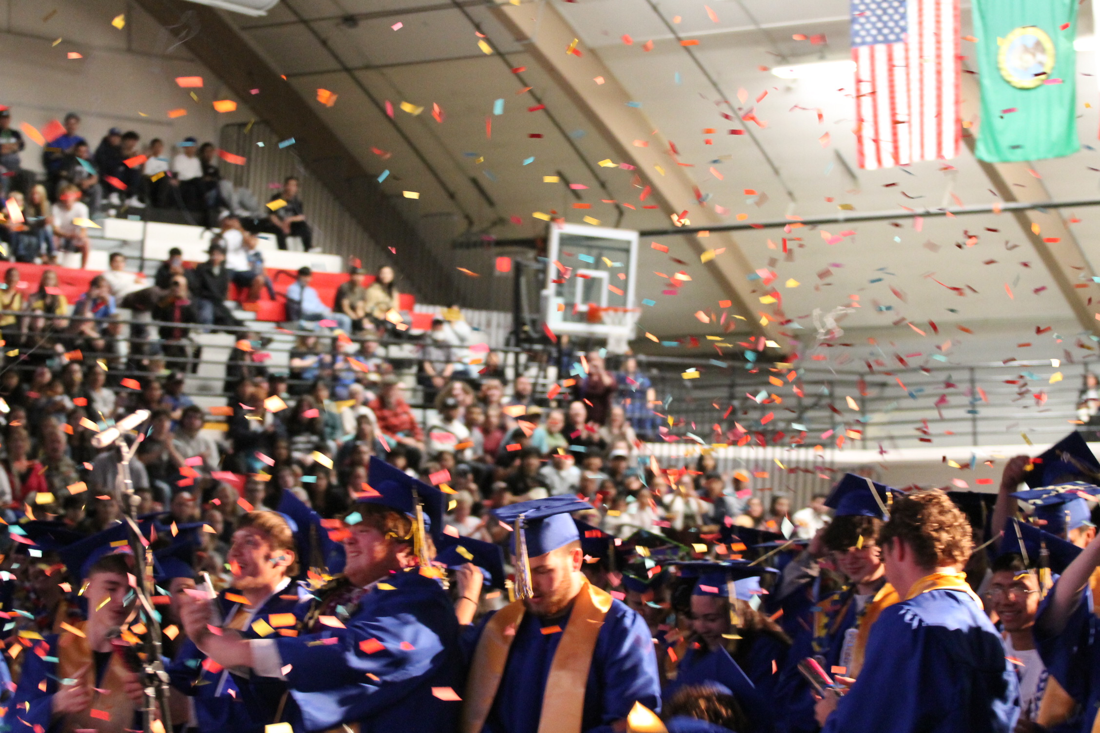 A graduation ceremony with graduates in blue gowns and caps. Confetti falling. An American and green flag on the ceiling.
