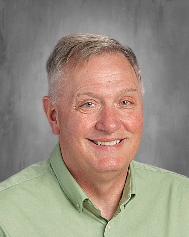 A man with short hair wearing a green shirt smiles at the camera. He is in front of a gray background.
