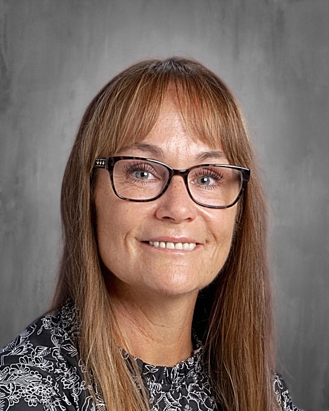 Woman with brown hair and glasses smiles at the camera. She is standing against a gray background.