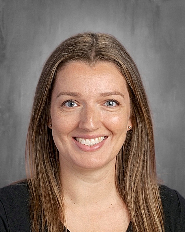 A woman with long brown hair smiles for a picture. She wears a black top. The background is plain gray.