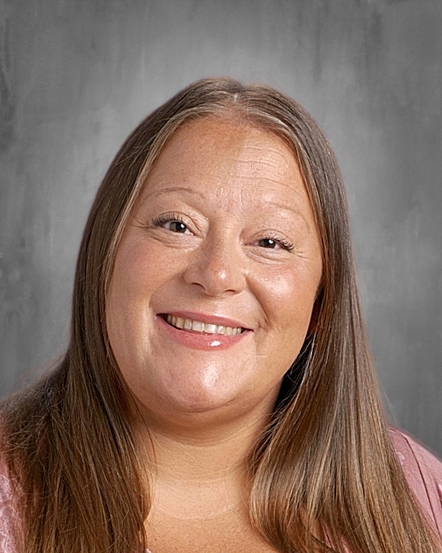 A woman with long brown hair smiling, wearing a pink top against a gray background.