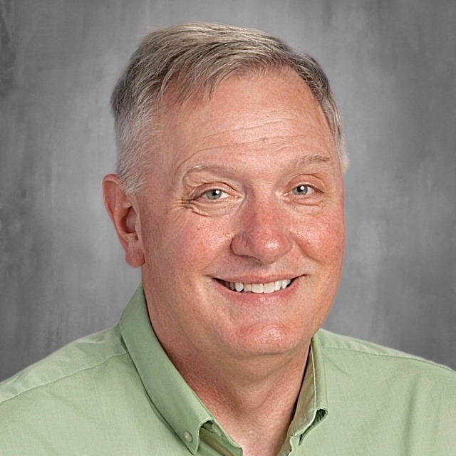 A man with graying hair, wearing a green shirt, smiling and looking at the camera.