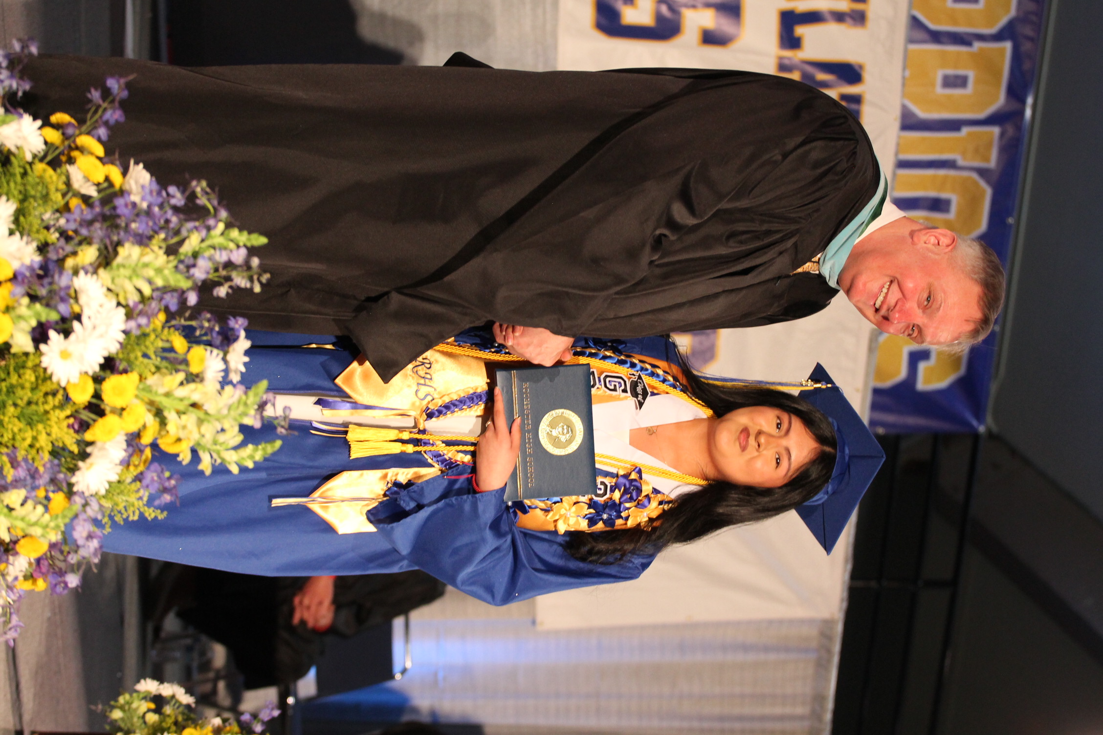 Man in black gown and woman in blue gown with cap present an award to a woman.