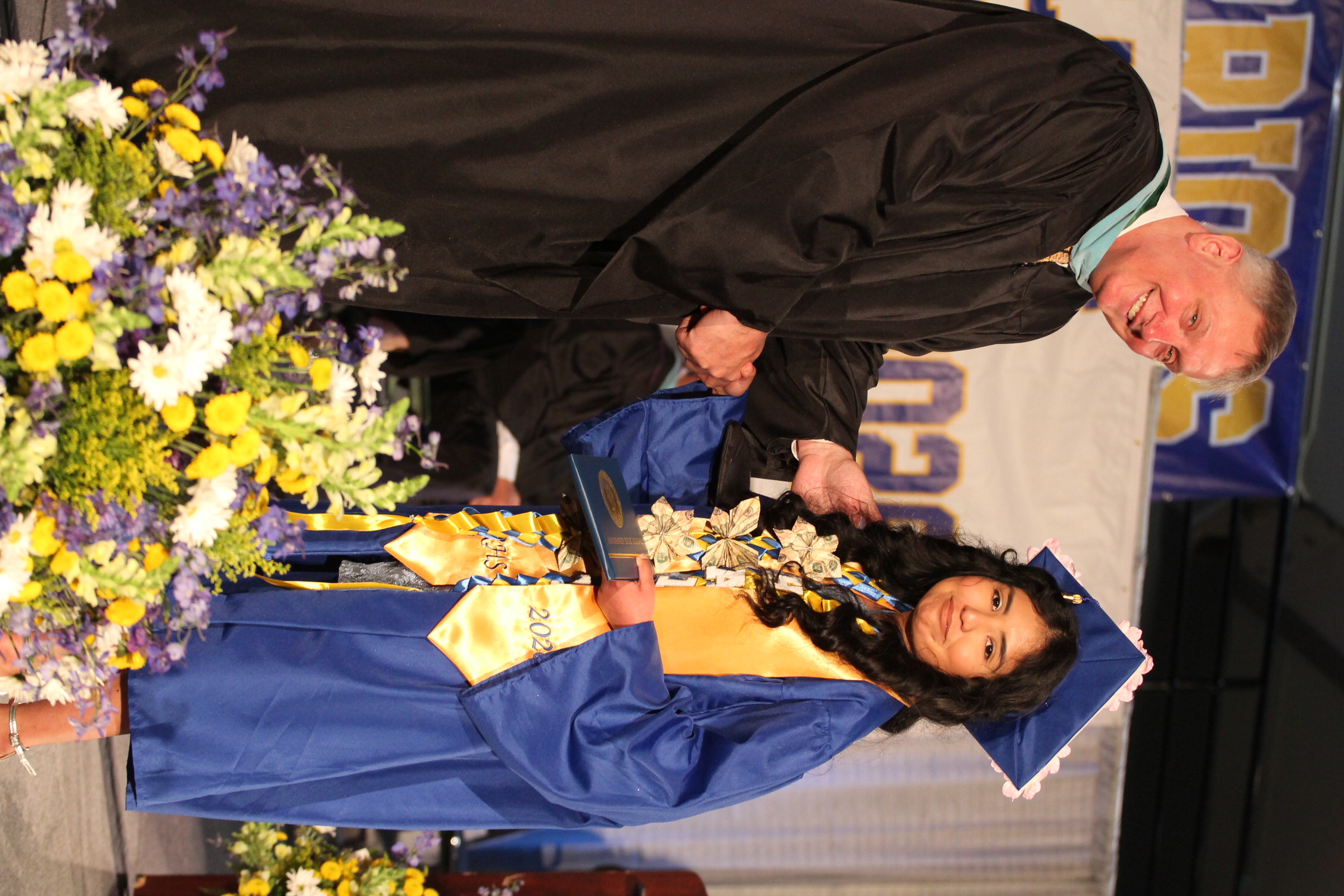 A man in a black robe shakes hands with a woman in a blue gown holding a trophy. They stand on a stage with flowers in the foreground.