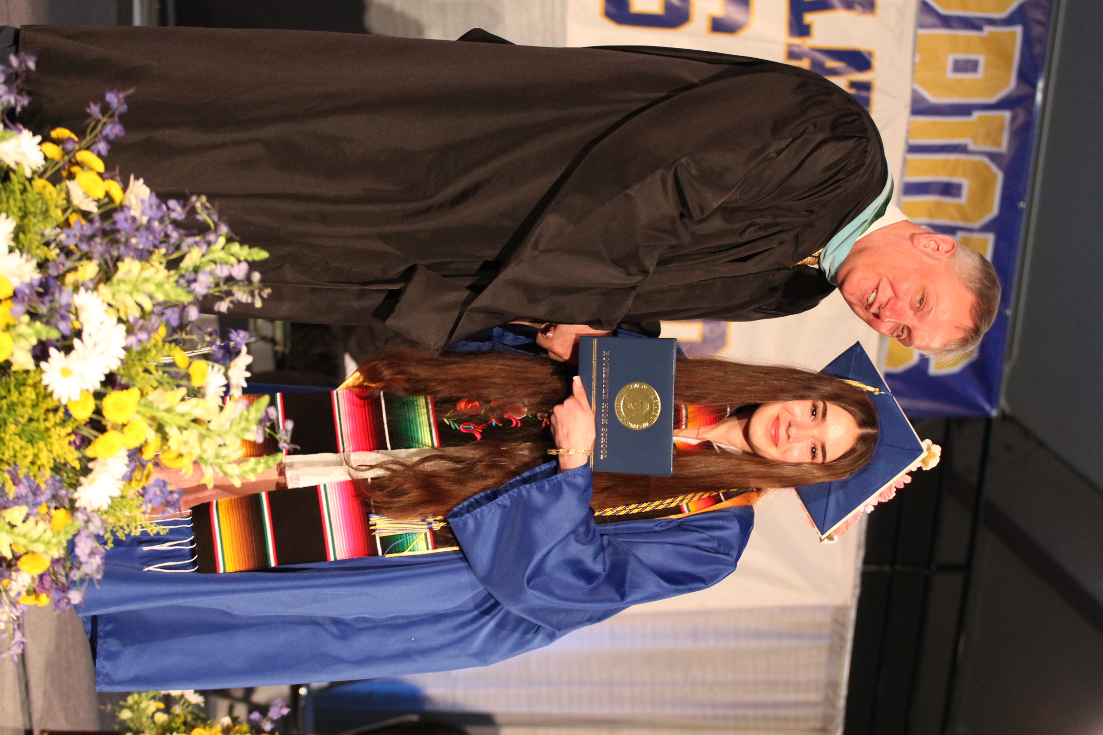 A man in a black robe hands a diploma to a young woman in a blue gown and cap.