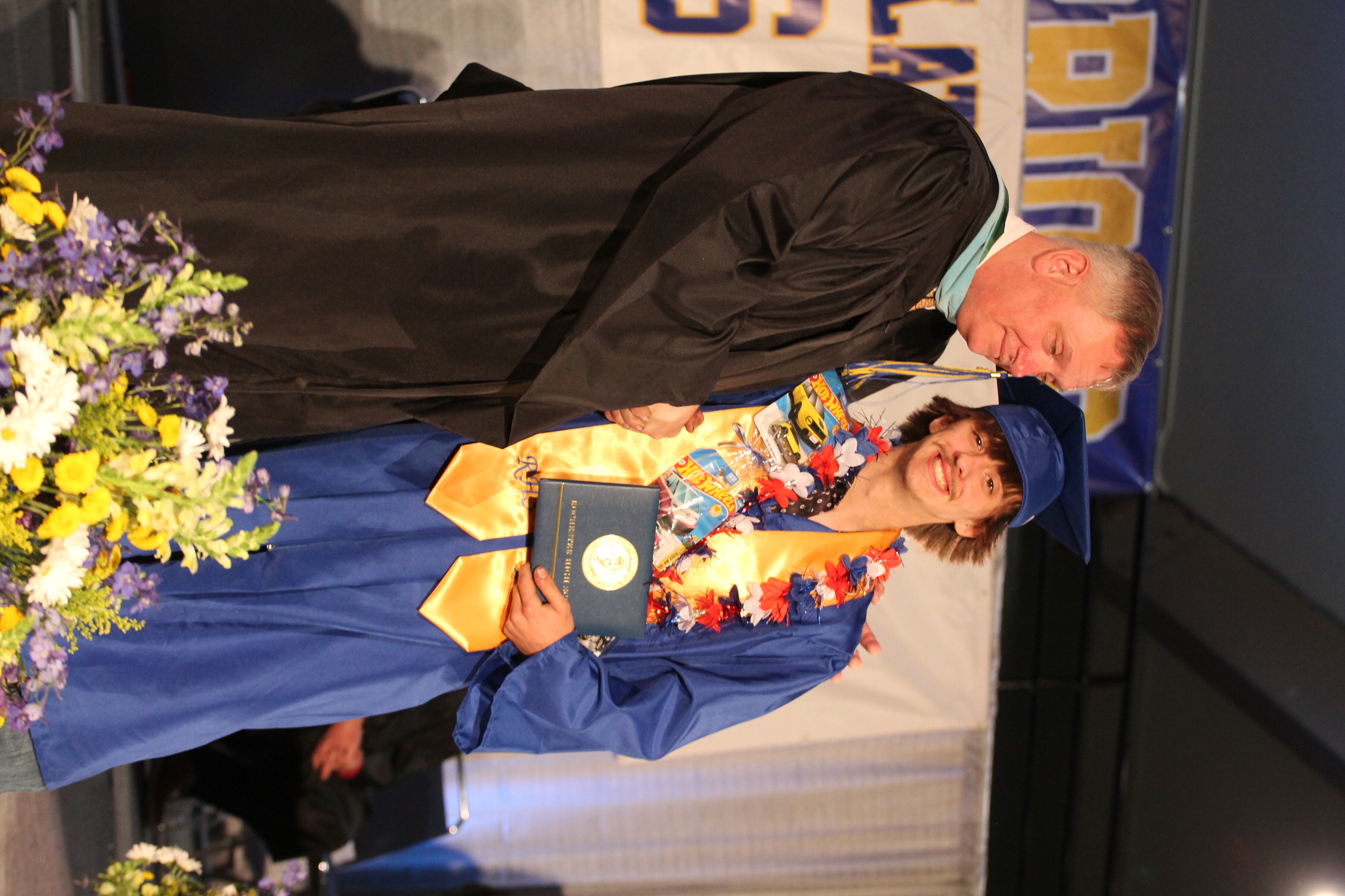 A man in a black graduation gown shakes hands with a graduate in blue, holding an award. They stand on a stage with flowers and a banner.