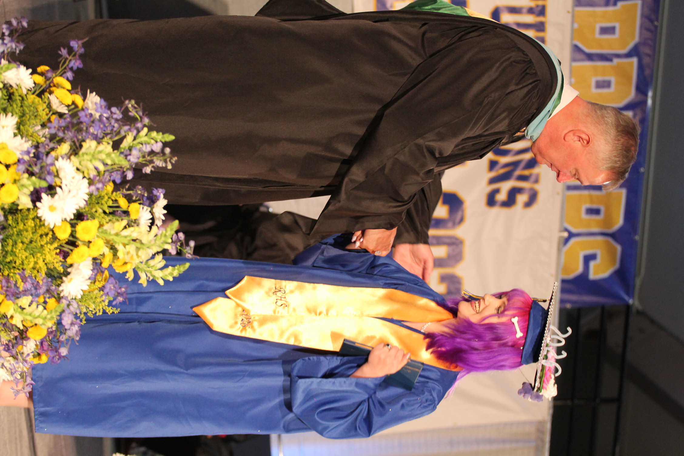 A man in a black graduation gown hands a diploma to a graduate wearing a blue gown and a colorful hat.