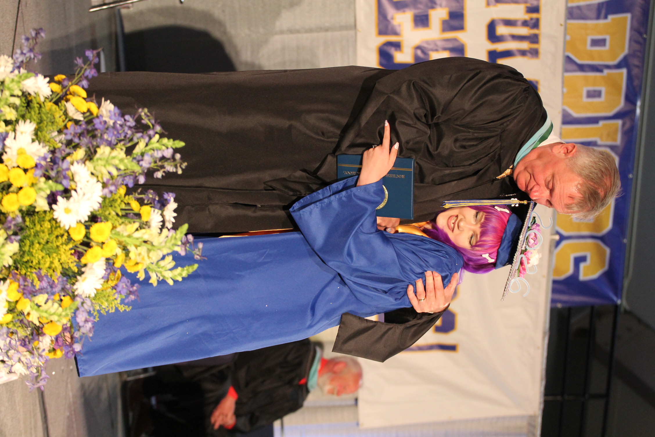A man and a woman in graduation attire on stage. The man hugs the woman, who holds a diploma. Flowers and banners are in the background.