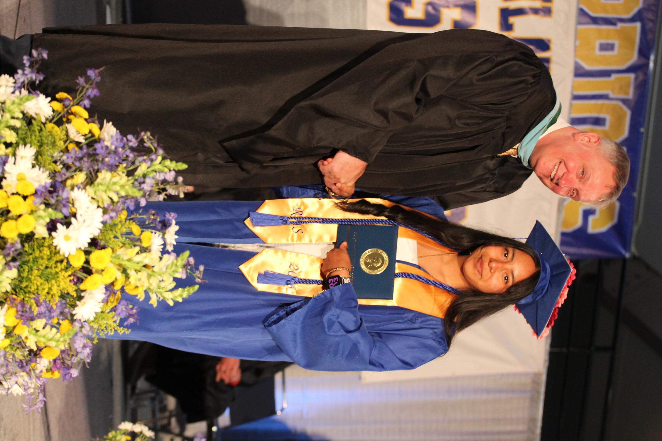 A male in a black gown hands a diploma to a woman in a blue gown. They stand beside a flower arrangement.