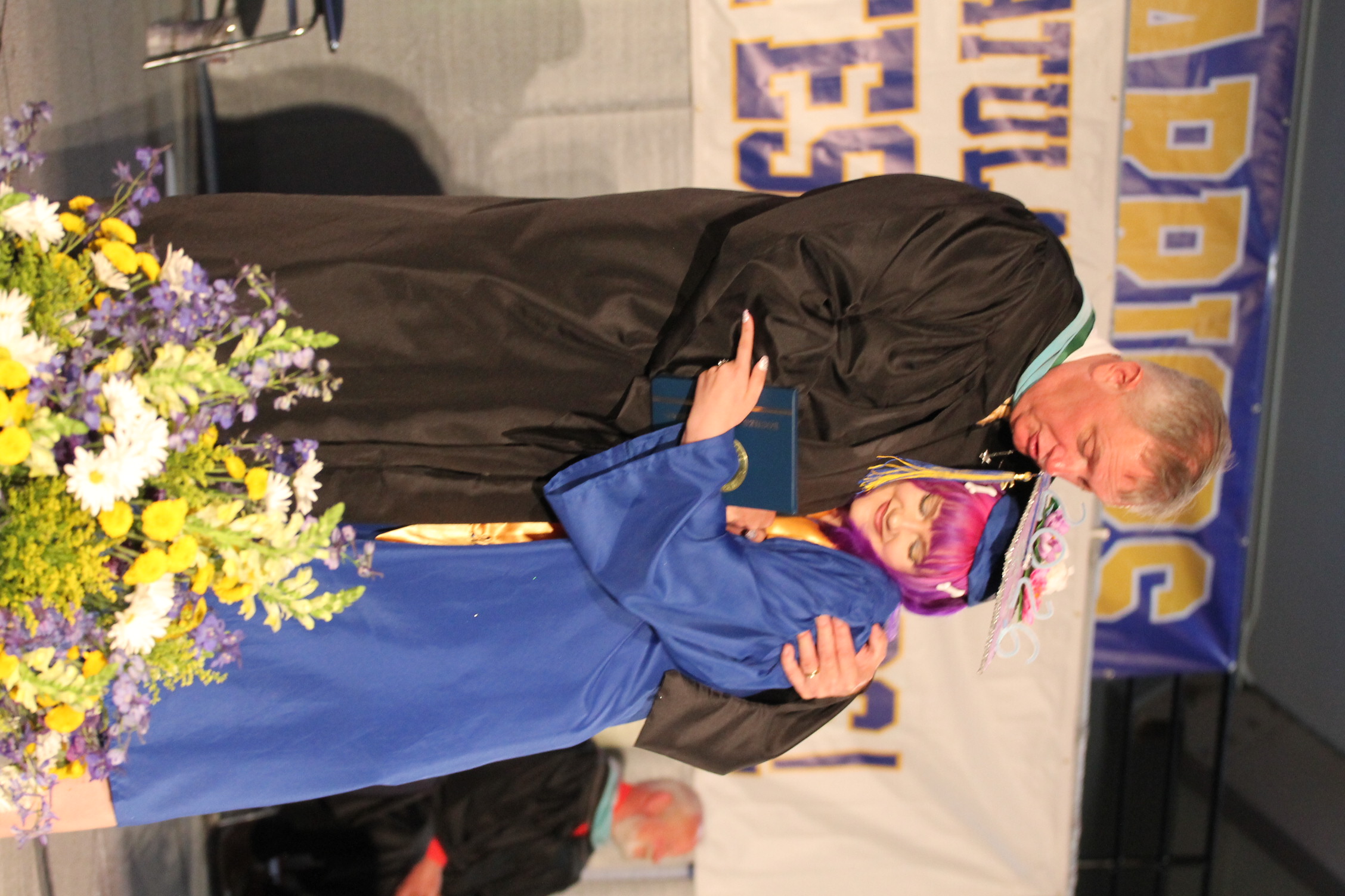 A male graduate advisor in a black robe congratulates a female graduate wearing a blue gown and purple wig.