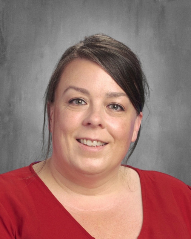 A woman with brown hair and a red shirt smiles at the camera against a gray background.
