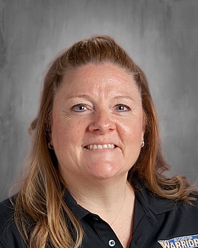 Woman with blonde hair wearing a black polo shirt with "WARRIOR" on it, smiling.