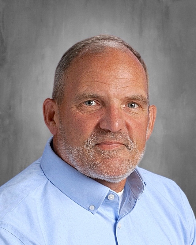 A man in a light blue shirt stands against a gray background, showing gray hair and a full beard.