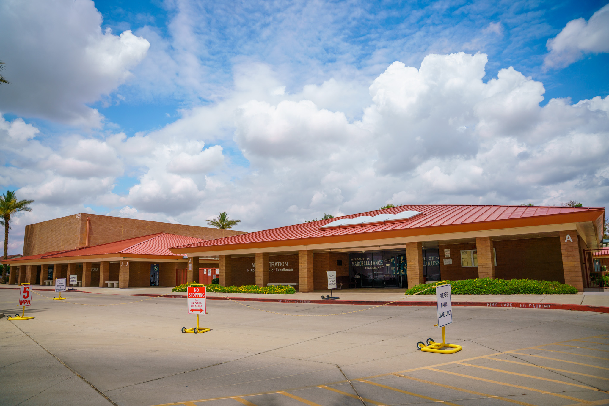 "The image shows the exterior of a school building with a red roof and beige brick walls. There are several signs in the parking lot, including one that reads '5 MPH,' another that says 'NO STOPPING,' and one that says 'PLEASE DRIVE CAREFULLY.' The building has large windows with text on them, including 'WELCOME TO MARSHALL RANCH ELEMENTARY' and 'ADMINISTRATION.' The sky is partly cloudy with patches of blue. There are palm trees visible in the background."