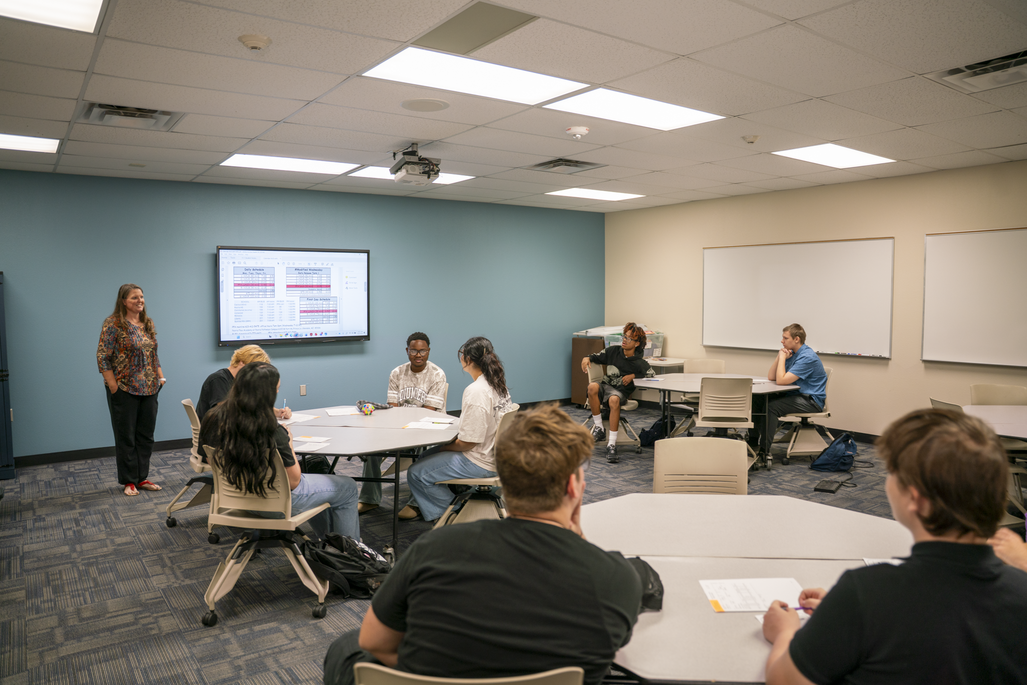 A classroom with students seated at round tables while an instructor stands at the front of the room beside a large digital display. The modern learning space features whiteboards, collaborative seating, and a projector mounted on the ceiling. Students are participating in a group discussion or lesson on the first day of class.