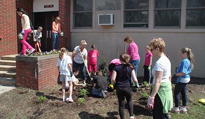 A group of children and adults are engaged in planting flowers and gardening outside a building, with some individuals working in the soil and others standing nearby.