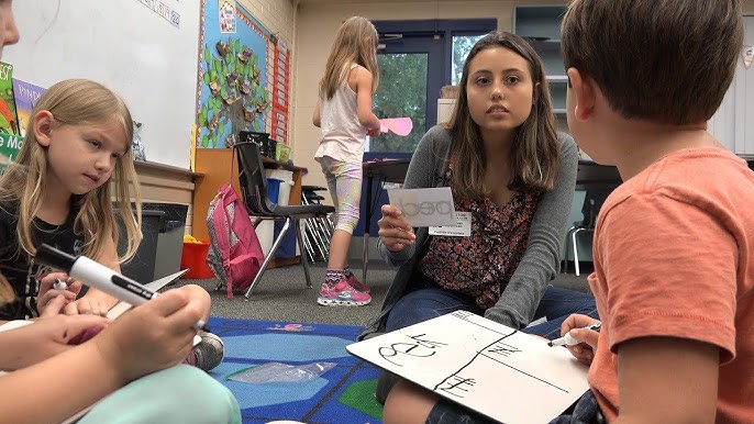 A teacher engages with a group of young students in a classroom setting, where one student is drawing on a whiteboard while others listen attentively.