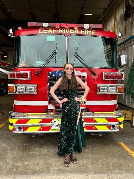 A girl in a green dress stands beside a bright red fire truck, smiling in a sunny outdoor setting.