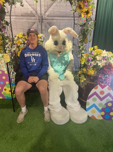 A man sits on a bench beside a large Easter bunny, creating a whimsical and festive atmosphere in the park.