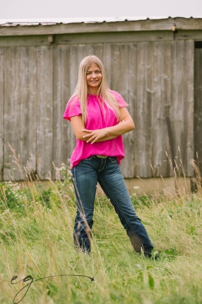 A blonde woman wearing jeans and a pink shirt stands confidently in front of a rustic barn.
