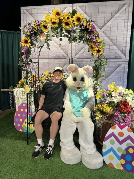A man seated on a chair beside a large Easter bunny, creating a whimsical and festive atmosphere.