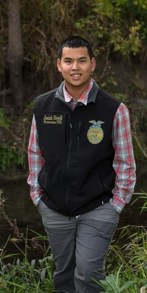 A young man in a vest stands beside a serene river, enjoying the natural surroundings and the calm atmosphere.