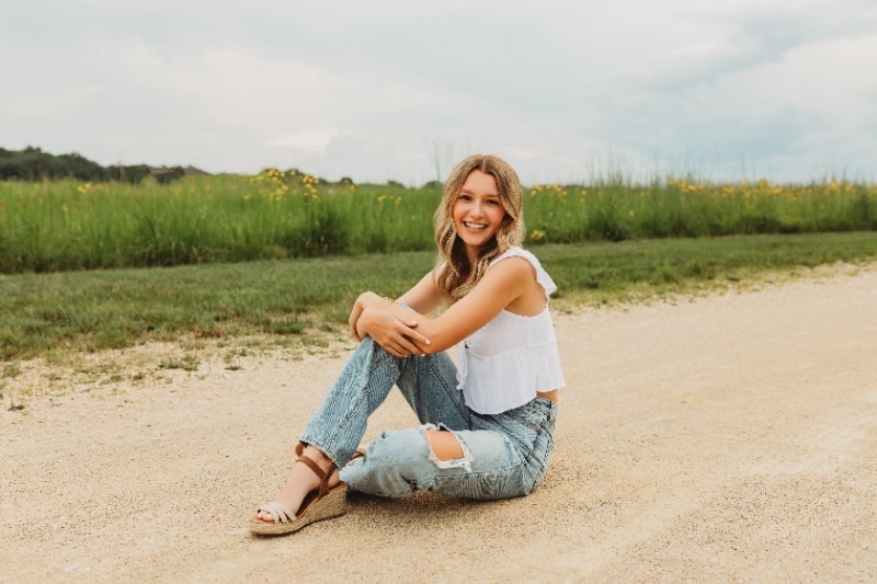 A smiling blonde woman in jeans and a white top sits on the road, exuding a cheerful and relaxed demeanor.