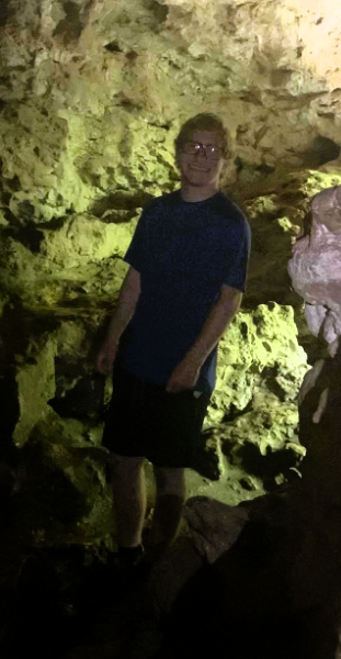 A man stands in a dim cave, illuminating the surroundings with a flashlight, revealing the rocky textures and shadows.