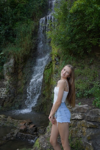 A girl in shorts stands beside a cascading waterfall, surrounded by lush greenery and sparkling water droplets.