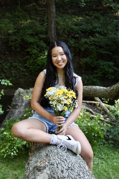 A girl sits on a log surrounded by colorful flowers, enjoying a serene moment in nature.
