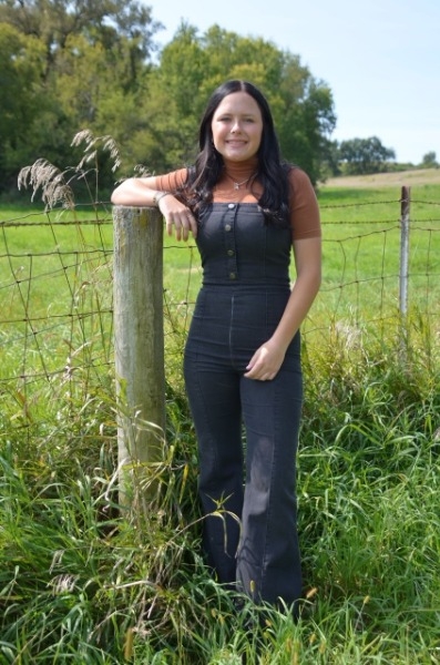 A woman in overalls stands beside a wooden fence, showcasing a casual and relaxed outdoor setting.