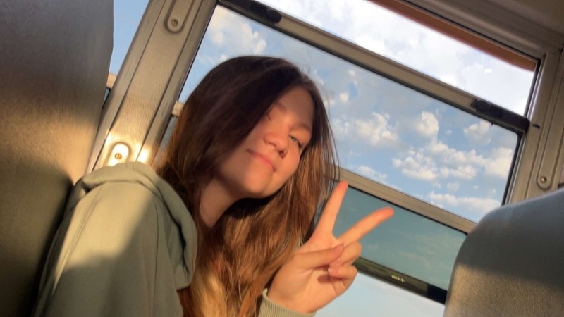 A girl sitting on a bus raises her hand, signaling for attention or assistance while looking out the window.