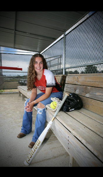 A young girl sits on a bench, holding a bat and ball, ready for a game in a sunny outdoor setting.