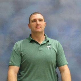 A man wearing a green shirt is seated on a chair, appearing relaxed and contemplative.