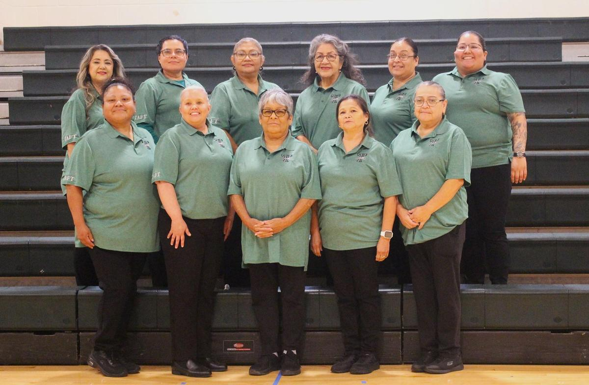 A group of women wearing green shirts stands together on bleachers, smiling and engaging with one another.