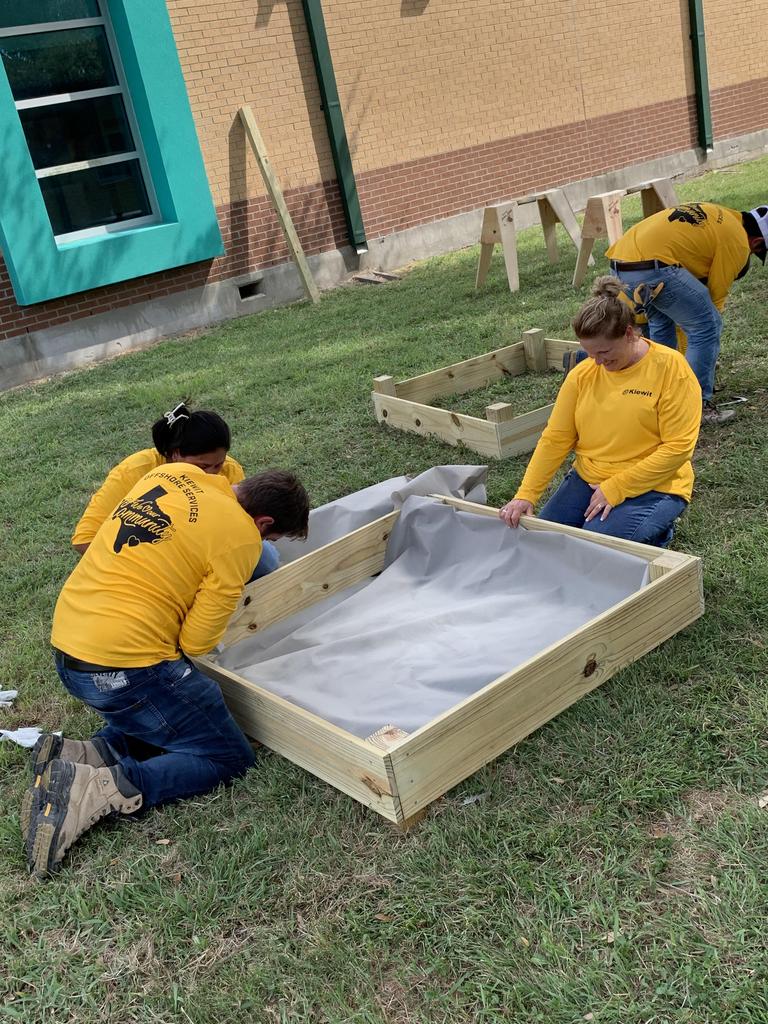 Three individuals in yellow shirts collaborate on constructing a raised garden bed outdoors.