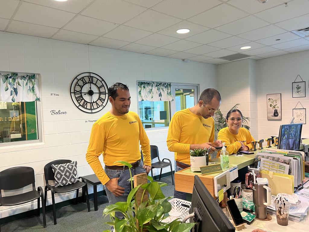 Three individuals wearing yellow shirts are standing together at a desk, engaged in a discussion or activity.