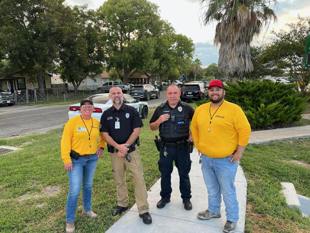 Three police officers stand on a sidewalk in front of a house, engaged in conversation and observing their surroundings.