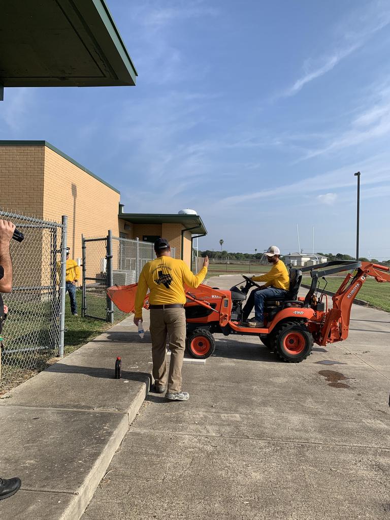 A group of individuals gathers around a tractor, with a man in a yellow shirt prominently standing among them.