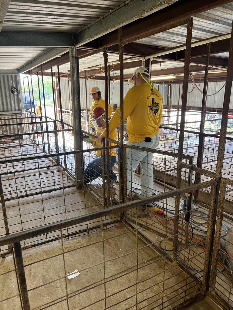 Two men in yellow shirts are constructing a cage inside a barn, focused on their task amidst the rustic surroundings.